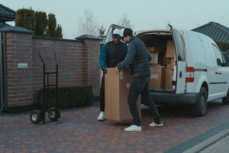 Two men engaged in a home relocation process are captured in the image, loading a large cardboard box into the back of a white delivery van parked on a brick driveway outside a residential property. T