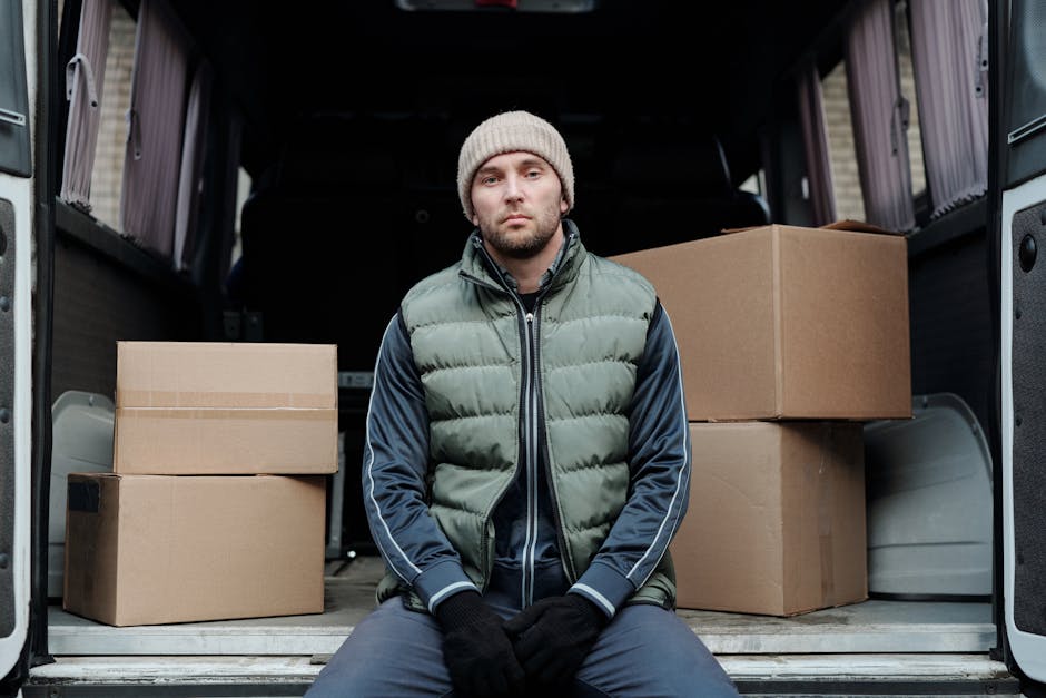 Two men engaged in a home relocation process are captured in the image, loading a large cardboard box into the back of a white delivery van parked on a brick driveway outside a residential property. T