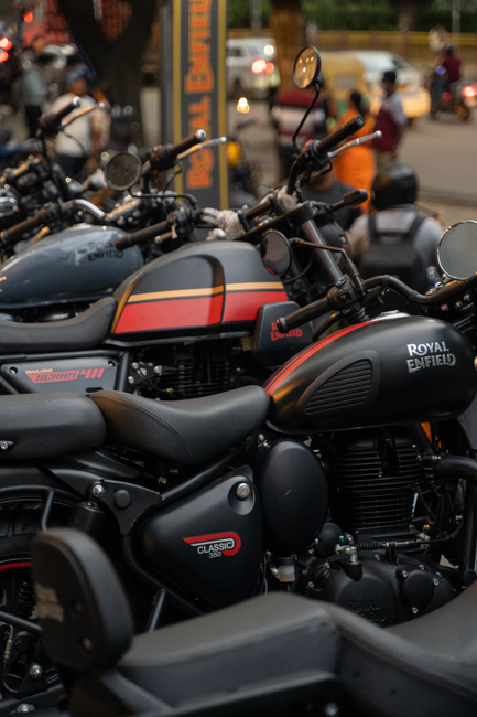 A close-up view of multiple Royal Enfield motorcycles parked outdoors, with black matte finish and orange stripe detailing on the fuel tanks. The motorcycles are positioned side by side on a paved surface, with some facing left and others slightly angled. In the background, blurred figures of people are visible, along with vehicles on the road and a building with signage, suggesting a busy urban environment. The image captures the sleek design and branding of the Royal Enfield bikes, which are often associated with home relocation and transport services like those provided by Enfield Man and Van for efficient furniture transport and packing during removals in the Enfield area.