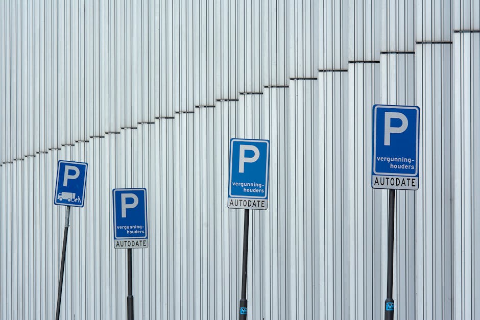 A rectangular white sign with bold black text reading 'NO PARKING DAY OR NIGHT' mounted on a grey garage door. The sign is attached with four metal screws, one in each corner. The garage door's surfac