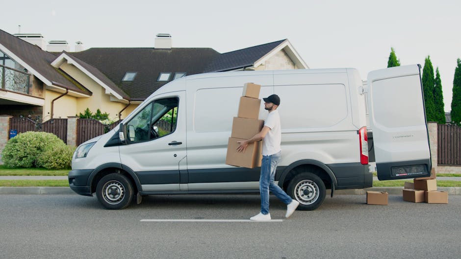 A man wearing a white t-shirt, blue jeans, and a black cap is in the process of loading large cardboard boxes into the side of a white panel van parked on a residential street. The van, used for home relocation and furniture transport, is positioned parallel to the curb, with the vehicle door open. Several smaller boxes are placed on the ground nearby on the pavement, some waiting to be loaded or unloaded. The background features modern detached houses with pitched roofs, chimneys, and well-maintained gardens enclosed by wooden fences, while tall green trees line the street. The scene is set during daylight with natural lighting, and the focal point is the loading process involving packing and moving activities supported by tools such as the boxes and the van, which is indicative of professional removals services offered by companies like Enfield Man and Van.