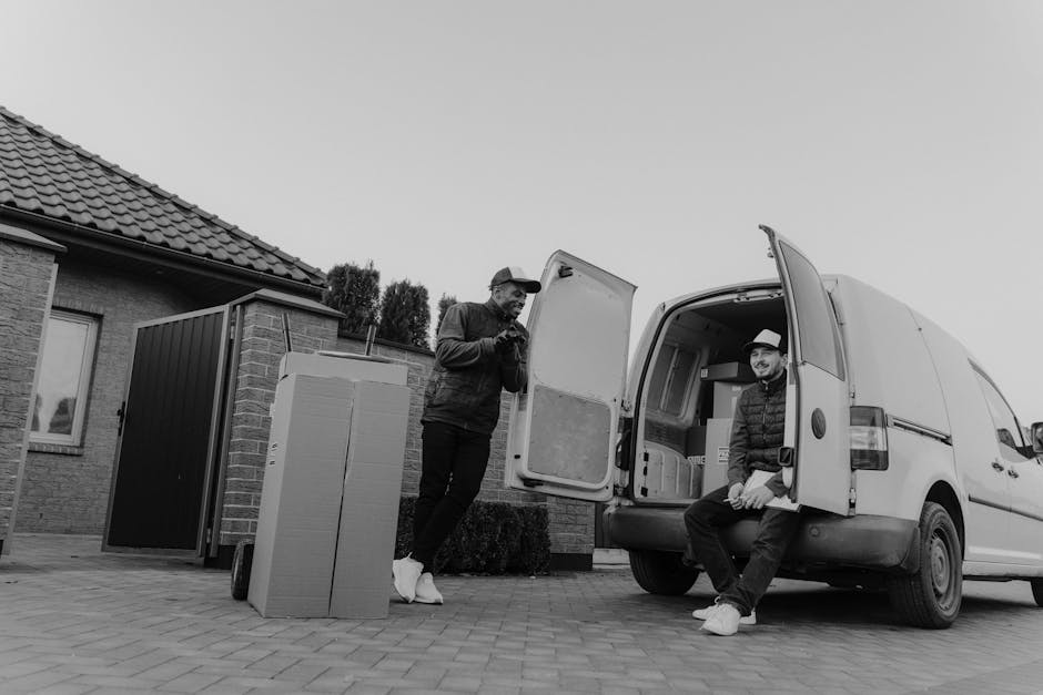 Two men involved in a home relocation process with a white moving van parked on a driveway in front of a residential property. One man, standing outside the van, is leaning on a large cardboard box with protective wrapping, likely used for packing furniture or household items. The second man is seated on the edge of the open rear door of the van, which is filled with various packed boxes and crates, some wrapped in plastic or fabric for protection. Nearby on the paved driveway, there is a large, flat-screen television still in its packaging, and a small trolley or cart is positioned close by, suggesting active loading or unloading. The house features brickwork and a tiled roof, with a window visible on the side wall. Bright natural light illuminates the scene, indicating it is daytime. This image illustrates the process of furniture transport, packing, and loading for house removals, relevant to services offered by Enfield Man and Van as part of professional moving solutions.