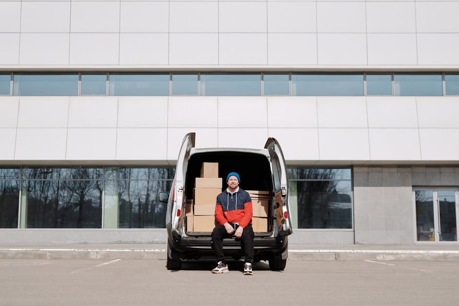 A man wearing a white t-shirt, blue jeans, and a black cap is in the process of loading large cardboard boxes into the side of a white panel van parked on a residential street. The van, used for home relocation and furniture transport, is positioned parallel to the curb, with the vehicle door open. Several smaller boxes are placed on the ground nearby on the pavement, some waiting to be loaded or unloaded. The background features modern detached houses with pitched roofs, chimneys, and well-maintained gardens enclosed by wooden fences, while tall green trees line the street. The scene is set during daylight with natural lighting, and the focal point is the loading process involving packing and moving activities supported by tools such as the boxes and the van, which is indicative of professional removals services offered by companies like Enfield Man and Van.