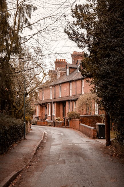 A narrow residential street leading to a row of three-story red-brick houses with pitched roofs, some featuring chimneys. The pavement is slightly wet, with a small curb on one side and a low brick wall bordering the front gardens. Tall, leafless trees and dark evergreen shrubs flank the street, partly obscuring the houses. A black wheelie bin is positioned near the curb, and a narrow alley runs alongside the houses. This scene depicts a quiet neighborhood environment suitable for home relocation activities. Enfield Man and Van, specializing in removals, often operate in similar settings where careful logistics are necessary for tight access areas, requiring precise loading of household furniture, boxes, and packing materials into a nearby van or moving truck, which is not visible in this image.