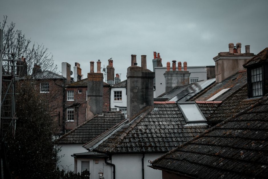 A view of a row of residential rooftops in a dense urban area during overcast weather, with multiple brick and tile roofs featuring chimneys and skylights. Some roofs are wet, indicating recent rain, and various chimney stacks with red brick and terracotta pots are visible among the rooftops. In the foreground, a white house with a dark, weathered tiled roof is seen, along with the edge of a wooden scaffolding structure on the left side. The background shows additional rooftops and chimneys, with muted sky overhead. This scene reflects typical UK home rooflines and chimney stacks, relevant to house removals and moving logistics, where careful handling of roof structures and access points is essential, as managed by services like Enfield Man and Van.