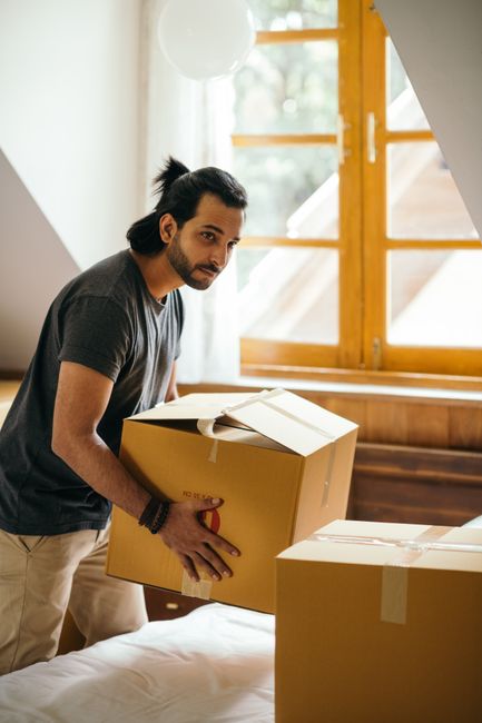 A man with dark hair and a beard, dressed in a dark grey t-shirt, is inside a residential room near a large wooden-framed window with open panes, natural light illuminating the space. He is lifting a sealed cardboard box, which appears to contain household items, while standing beside several other packed boxes of varying sizes stacked on a bed. The room has a wooden floor and light-colored walls, with visible household furniture and packing materials. This scene depicts the initial stages of home relocation, involving packing and preparation for furniture transport, as part of a professional removals process by Enfield Man and Van, specializing in house removals and moving services in the Enfield area.
