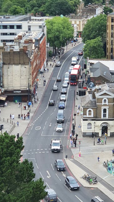 An aerial view of a residential street during daytime, showing multiple vehicles including cars, vans, and a double-decker bus travelling along a curved road, with some vehicles moving and others parked along the curb. The street is lined with a mix of historic and modern buildings, with pedestrians walking on the sidewalks and crossing at designated crossings. Several bicycles are parked near a small building at the corner, and green trees provide shade along the street. The scene captures an active urban environment, typical of an area where home relocation and furniture transport services like those offered by Enfield Man and Van may be required, with the clear visibility of traffic flow and pedestrian activity supporting efficient moving logistics and packing processes.