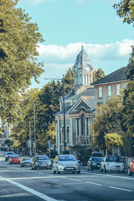 A street scene showing several parked cars along the kerbside on a residential road in Enfield, with a historic clock tower church featuring a tall spire in the background, surrounded by large leafy trees and residential buildings with brick facades. In the foreground, a property with an open doorway reveals a home relocation process involving the loading of furniture and packed cardboard boxes onto a van operated by Enfield Man and Van, a professional removals service. The scene captures the outdoor environment during daylight hours, with clear skies and natural sunlight illuminating the street and objects, highlighting the practical aspects of furniture transport and packing during a house move, emphasizing the careful loading process typical of professional removals in Enfield.